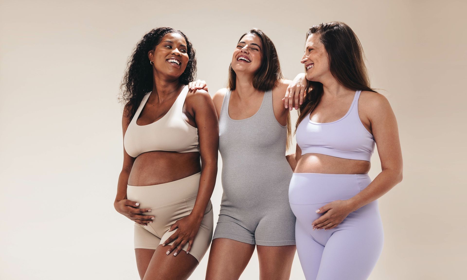 Three pregnant women in athletic wear smiling and holding their bellies.