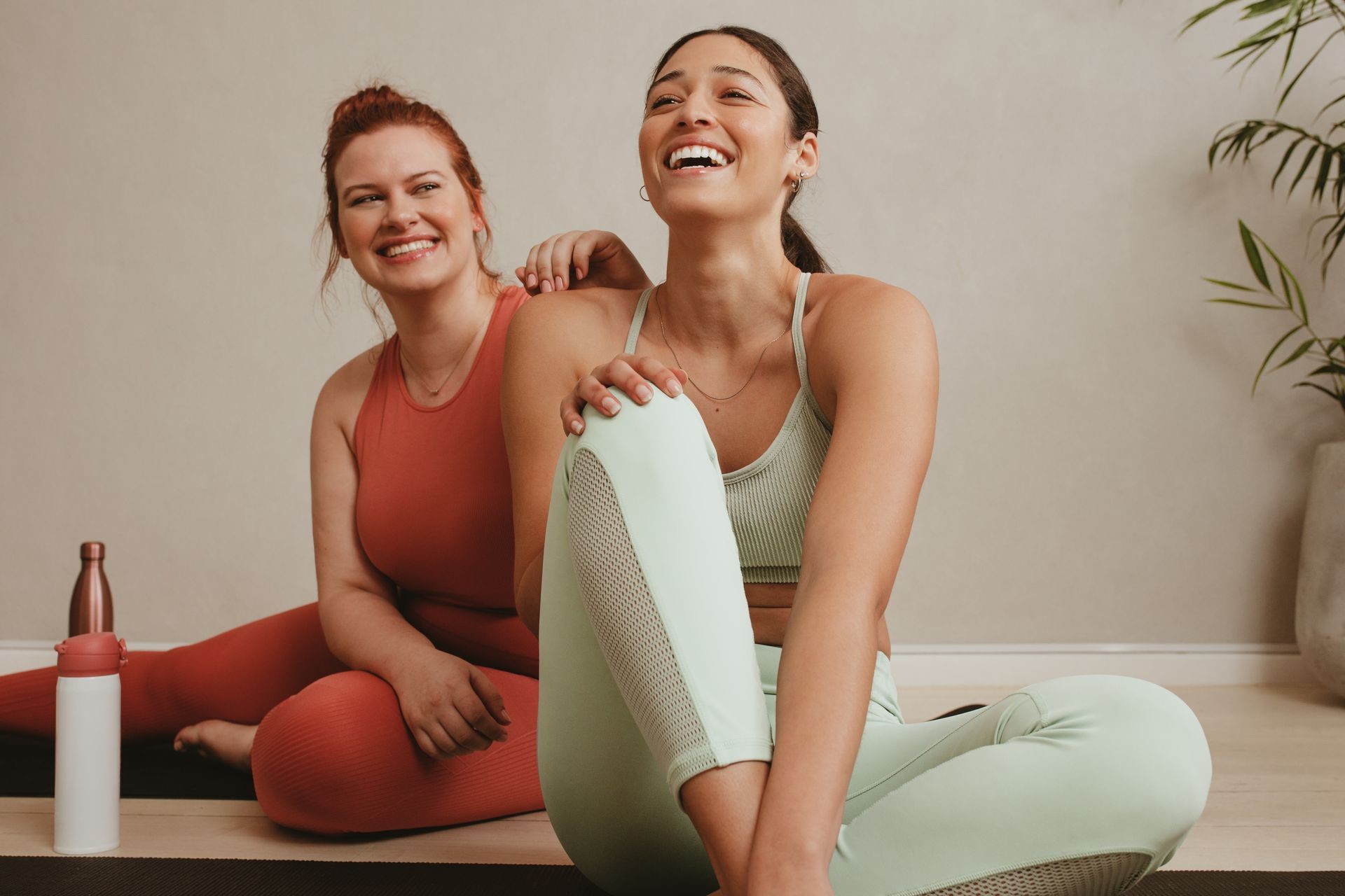 Two women in yoga attire sitting on mats, with a water bottle and plant in the background.