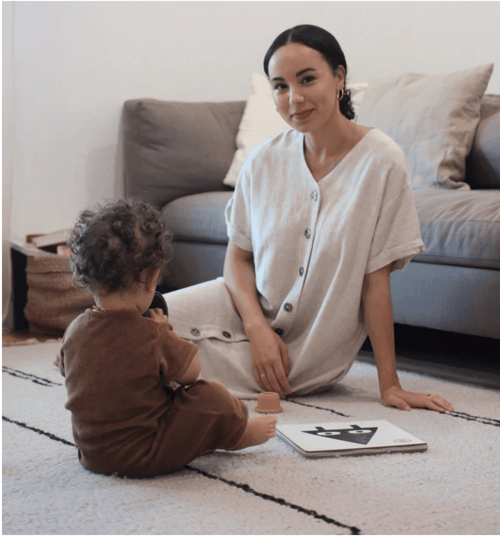 Woman sitting on a rug with a toddler playing, near a gray couch and pillows.