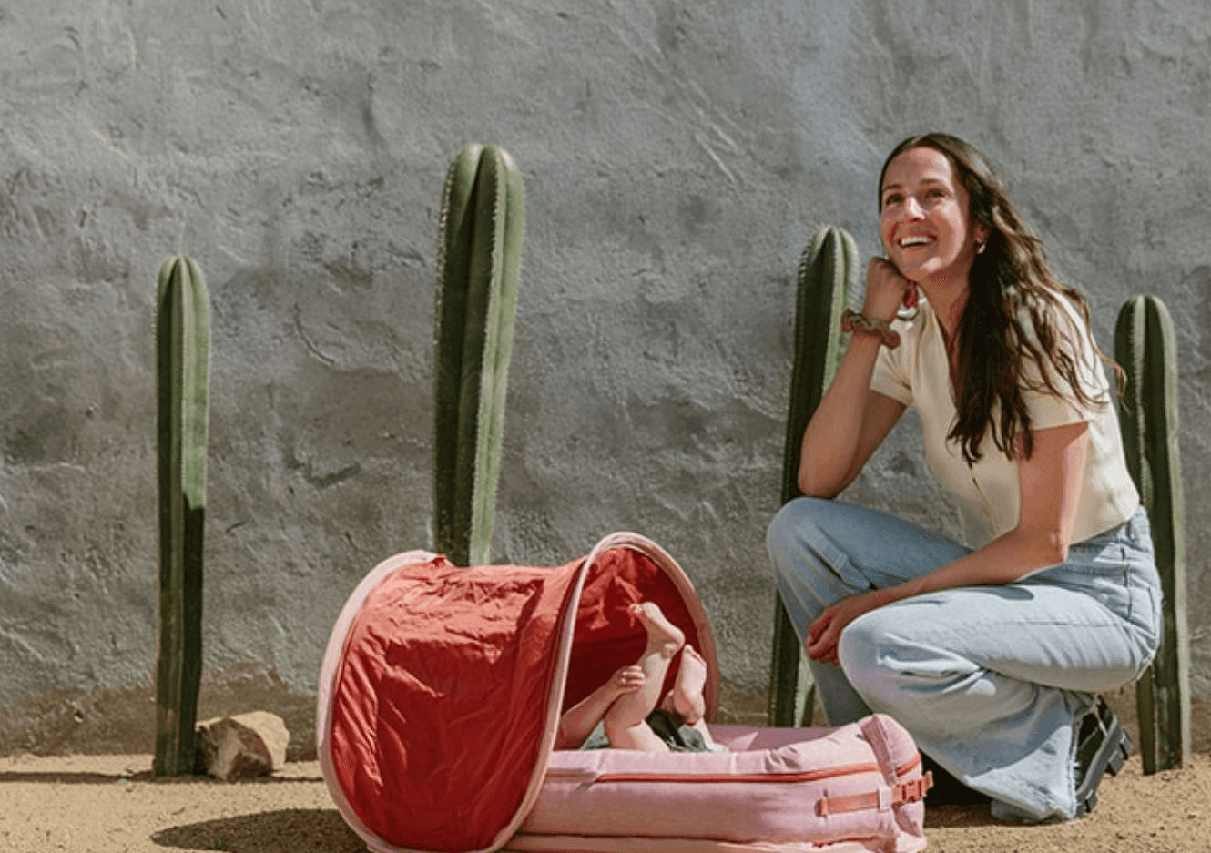 Woman smiling outdoors next to a baby in a pink tent with cacti in the background.