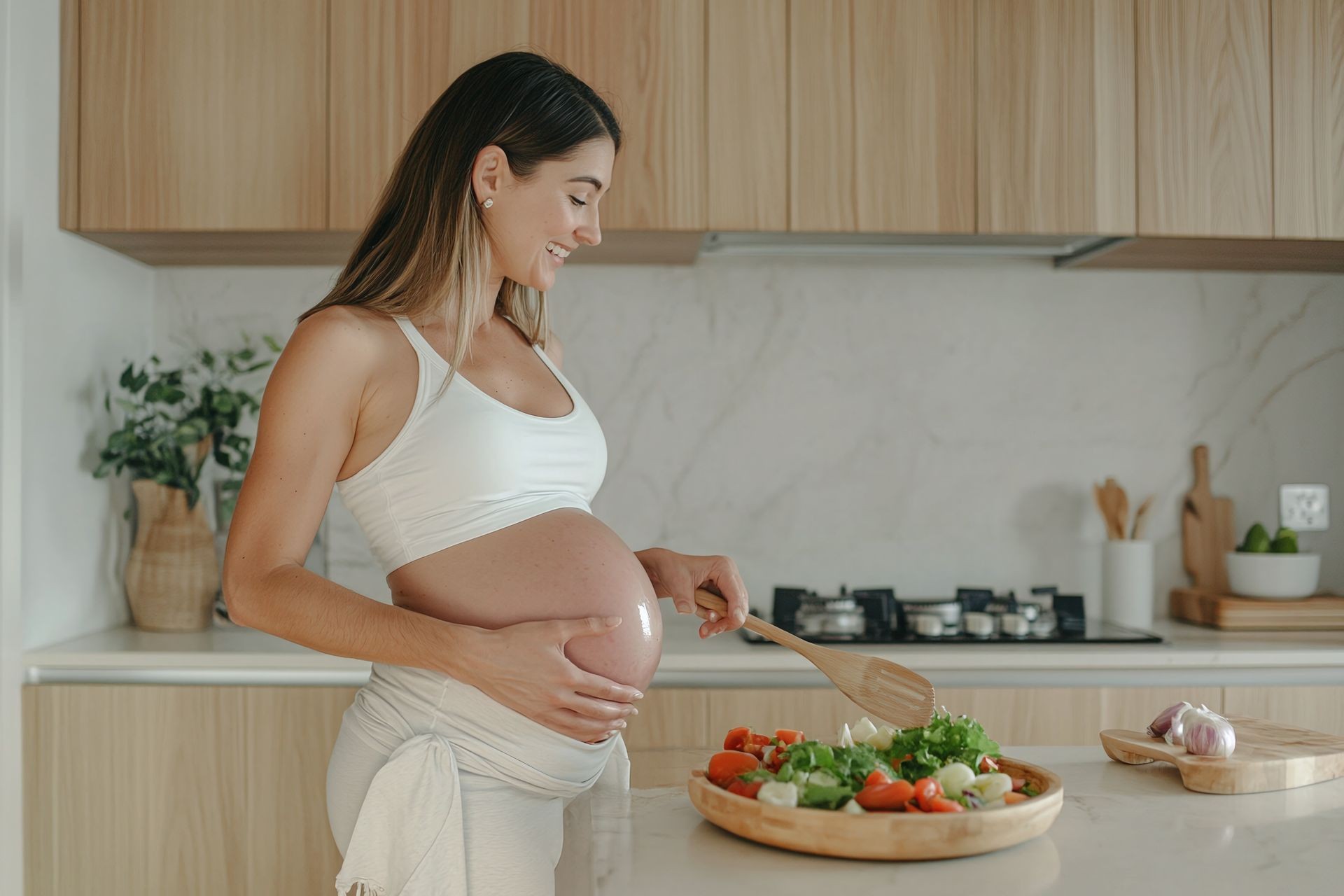 Pregnant woman in a white outfit preparing salad in a modern kitchen.