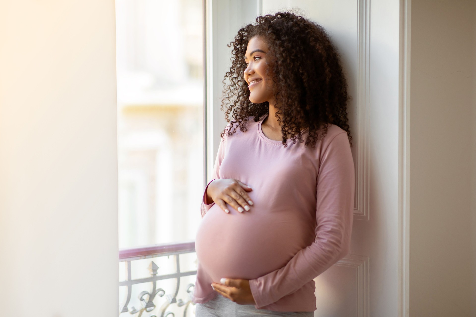 Black pregnant woman gently cradling belly while standing by window at home